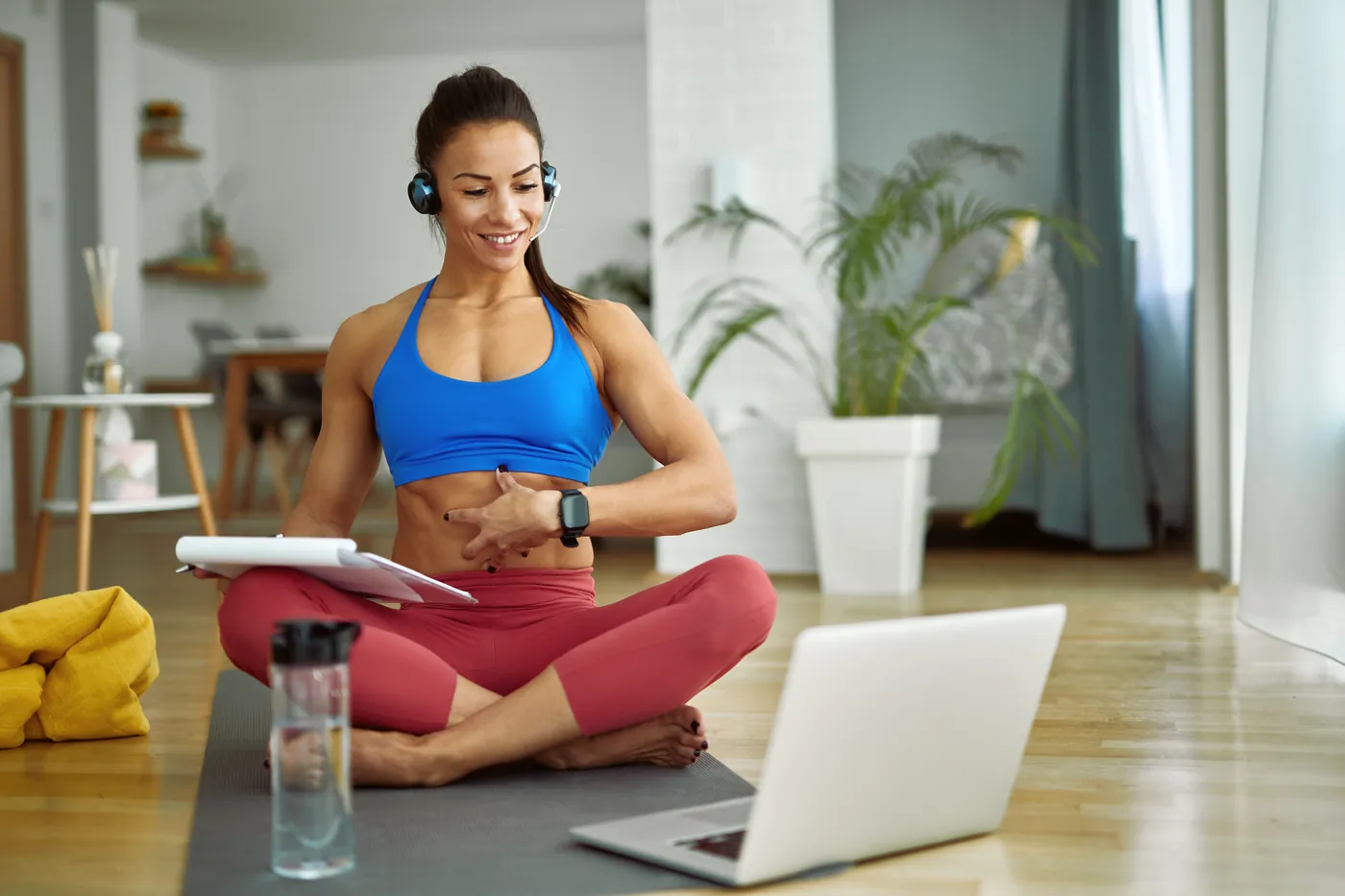Aluna sentada no tapete, com fone e laptop, conversa por vídeo antes do treino em casa.